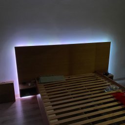 Starz-Drew - Drewniana rama łóżka z podświetleniem LED, widoczne szafki nocne z akcesoriami i butelka wody na stelażu łóżka.