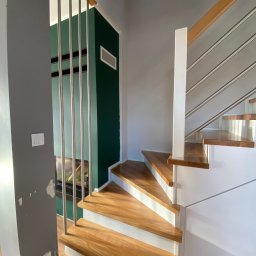 Starz-Drew - Nowoczesne schody z drewnianymi stopniami, białymi podstopnicami i metalowymi balustradami, widok z dołu, ściana w tle szara z widocznymi śladami po naprawach, zielona ściana w tle.