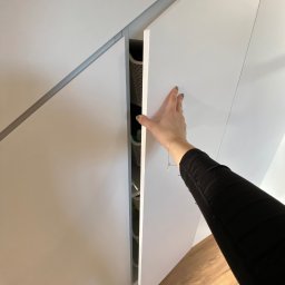 Starz-Drew - Biała szafa wnękowa z szufladami, częściowo otwarta przez kobiecą dłoń z bransoletką, widoczny parkiet na podłodze.