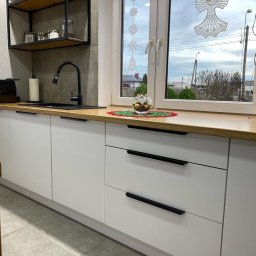 Starz-Drew - Nowoczesna kuchnia z białymi frontami szafek i drewnianym blatem, czarny zlew i bateria, okno z dekoracjami świątecznymi, widok na domy za oknem.