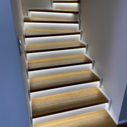 Starz-Drew - Nowoczesne schody z drewnianymi stopniami i białymi podstopnicami, podświetlone listwami LED, zrealizowane w Śmiglu. Widok z dołu, podkreślający minimalistyczny design i ciepłe światło.