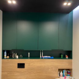 Starz-Drew - Zielone szafki łazienkowe podwieszane, z otwartą półką na kosmetyki i akcesoria, drewnopodobna zabudowa z ukrytym stelażem WC, białe płytki na ścianach, czarny sufit z oświetleniem punktowym.