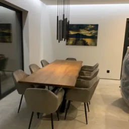 Stół z blatem z litego drewna o nieregularnych krawędziach, otoczony krzesłami tapicerowanymi w odcieniach szarości, oświetlony nowoczesną lampą wiszącą na tle minimalistycznego wnętrza z obrazem.