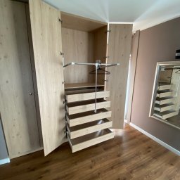 Starz-Drew - Otwarta szafa wnękowa z jasnego drewna z wysuwanymi szufladami i drążkiem na wieszaki, widoczne zawiasy, brązowa podłoga i ściana w tle.