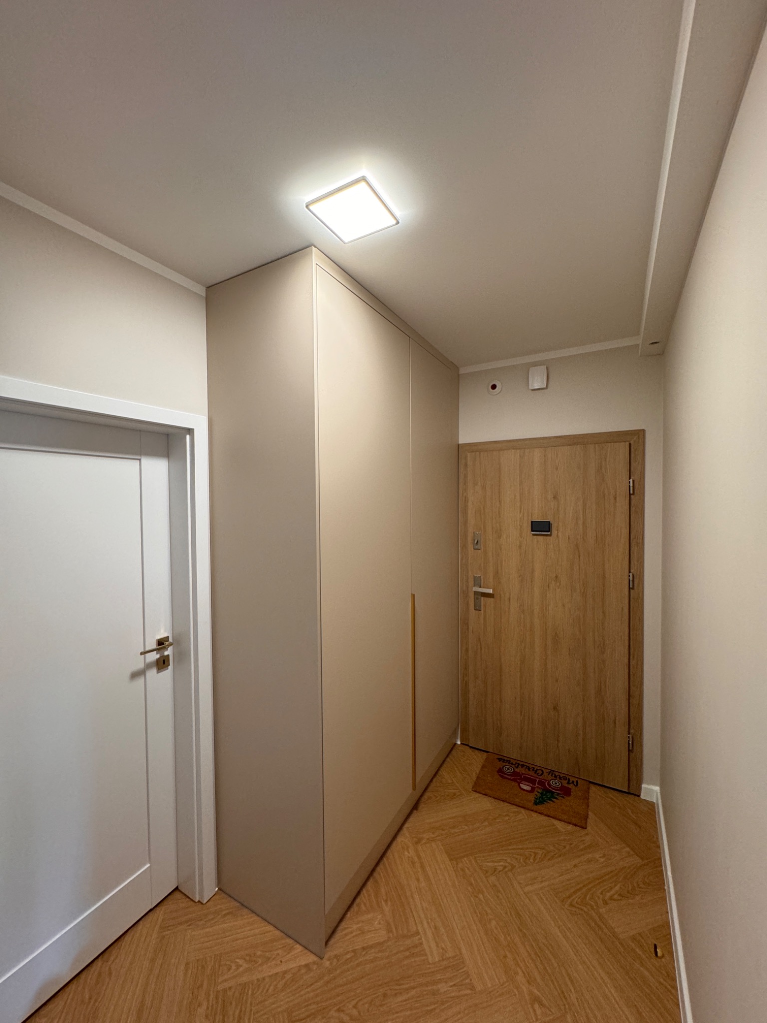 Beżowa szafa wnękowa na wymiar w korytarzu w Śmiglu, z drewnianą podłogą w jodełkę i świąteczną wycieraczką przed drzwiami wejściowymi.