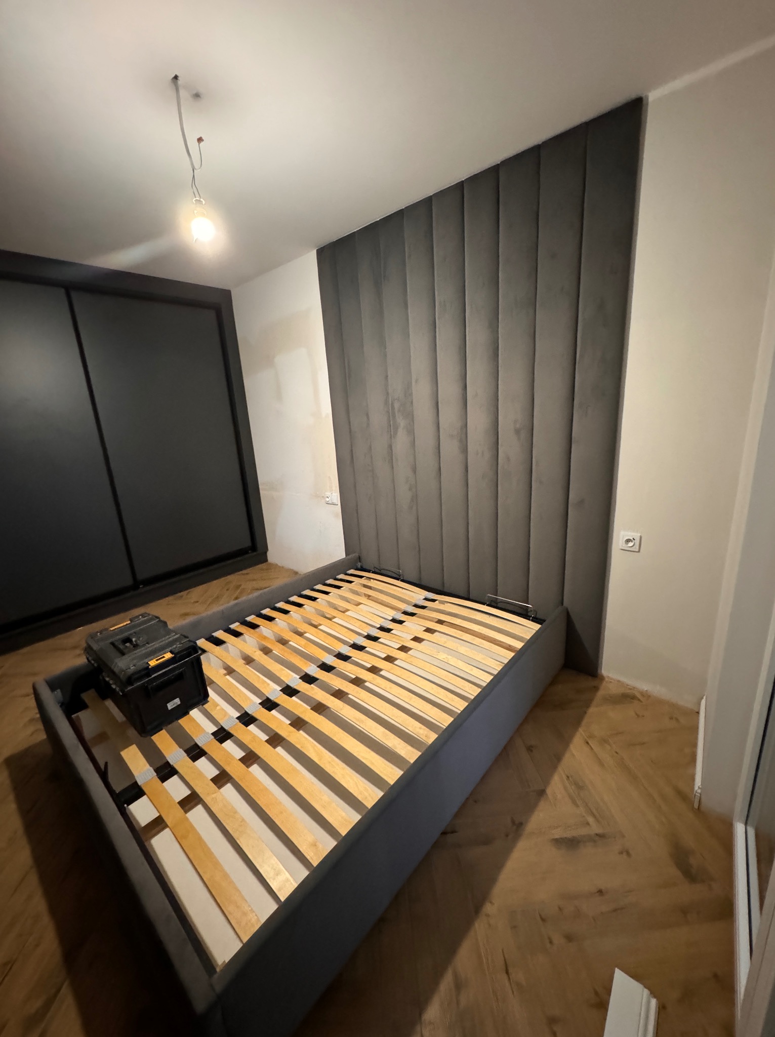 Szare tapicerowane łóżko z drewnianym stelażem, czarna szafa przesuwna w tle, skrzynka narzędziowa na ramie łóżka, parkiet ułożony w jodełkę, surowy stan wykończenia wnętrza, wisząca żarówka.