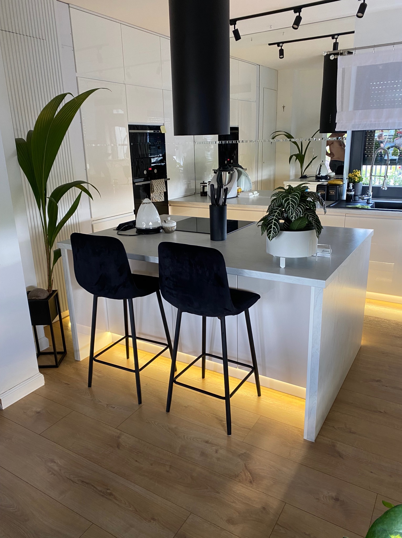 Wnętrze nowoczesnej, jasnej kuchni z wyspą, czarnymi hokerami i podświetleniem LED pod szafkami, widok na sprzęt AGD w zabudowie.