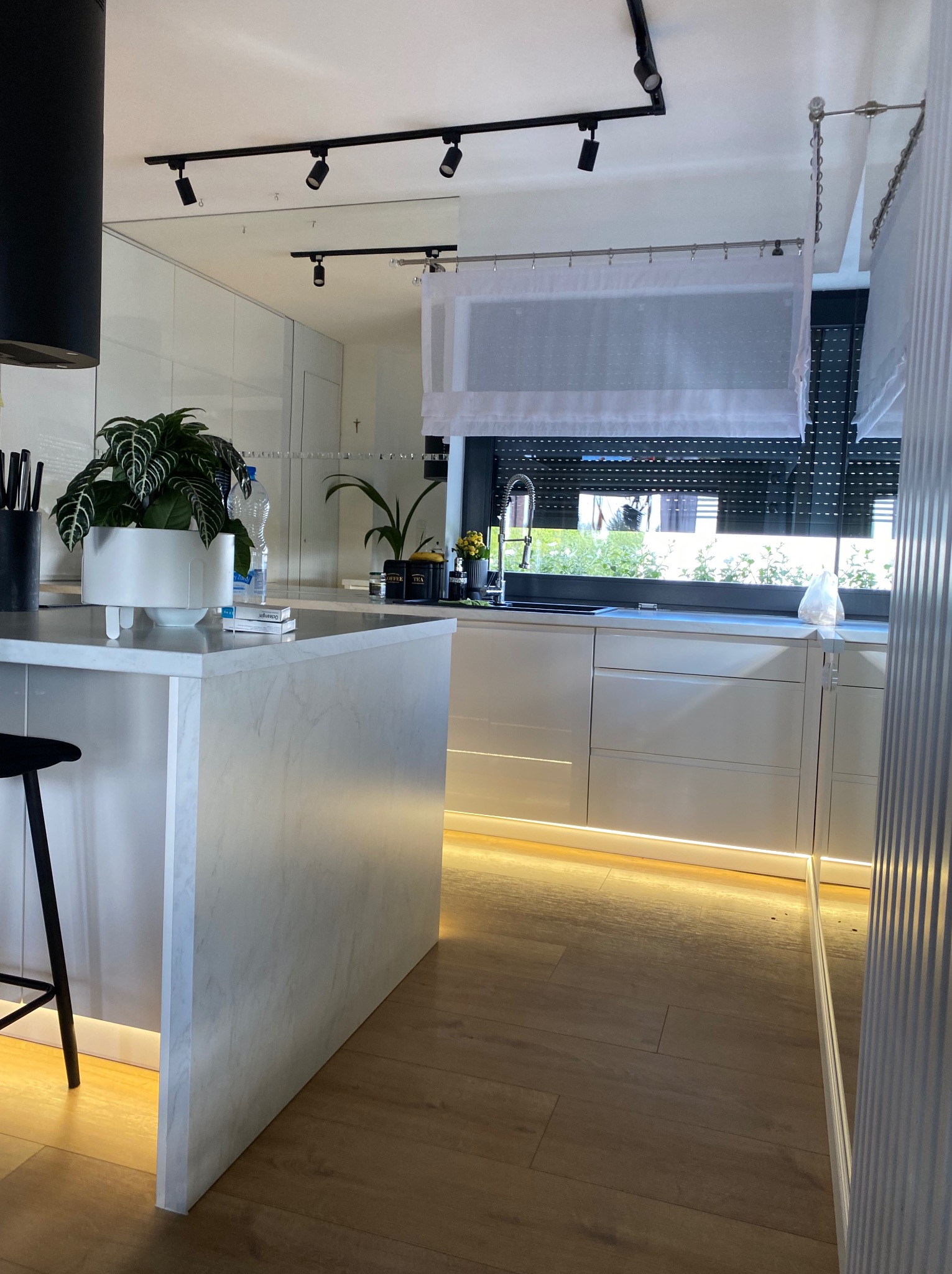 Nowoczesna, jasna kuchnia z wyspą wykończoną marmurem, oświetleniem LED pod szafkami i roślinami doniczkowymi na blacie, widok na okno z roletami i firanką.