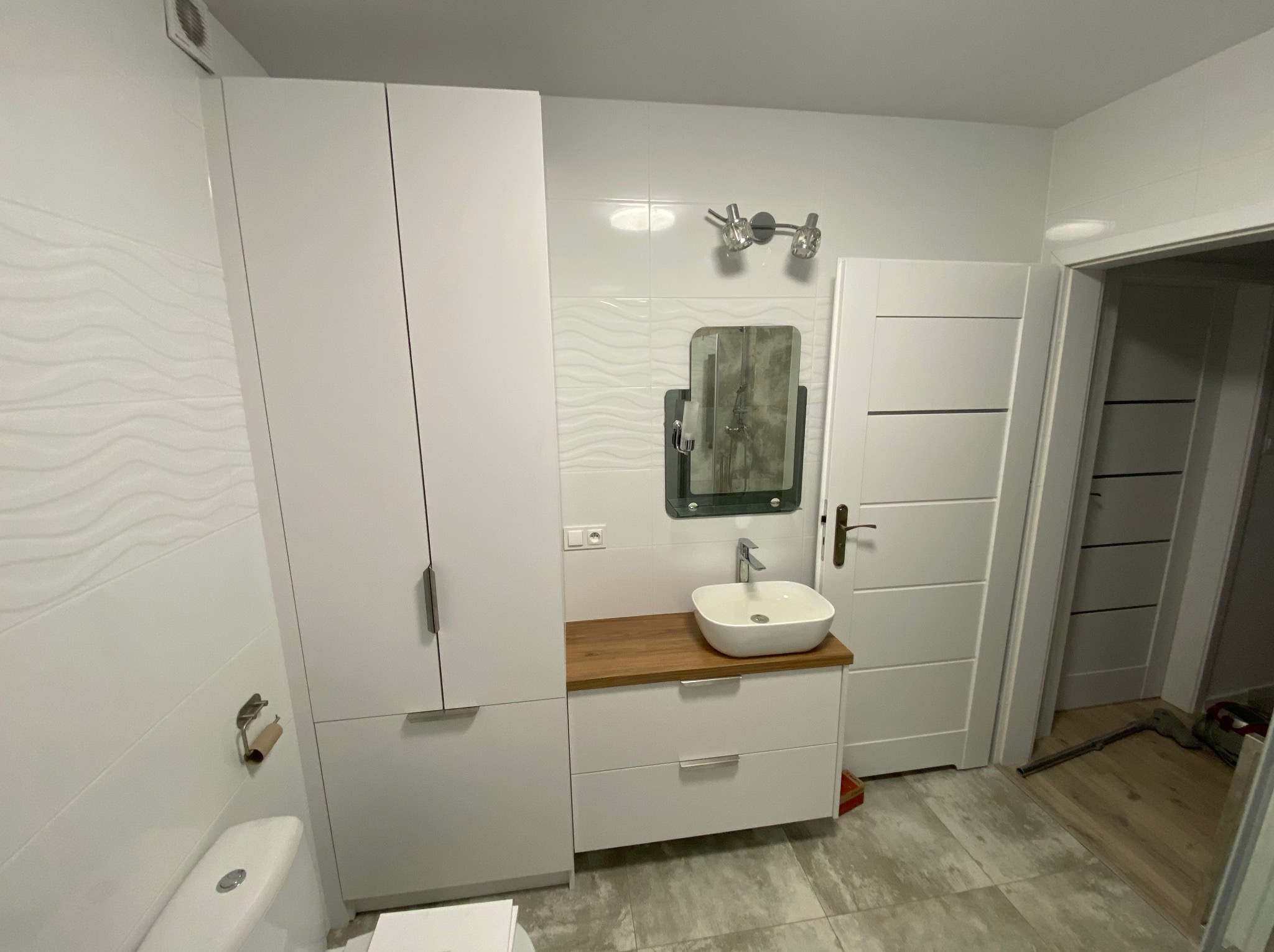 Nowoczesna łazienka z białą szafką pod umywalką z drewnianym blatem, lustrem w czarnej ramie, białą szafą i płytkami z subtelnym wzorem fal.