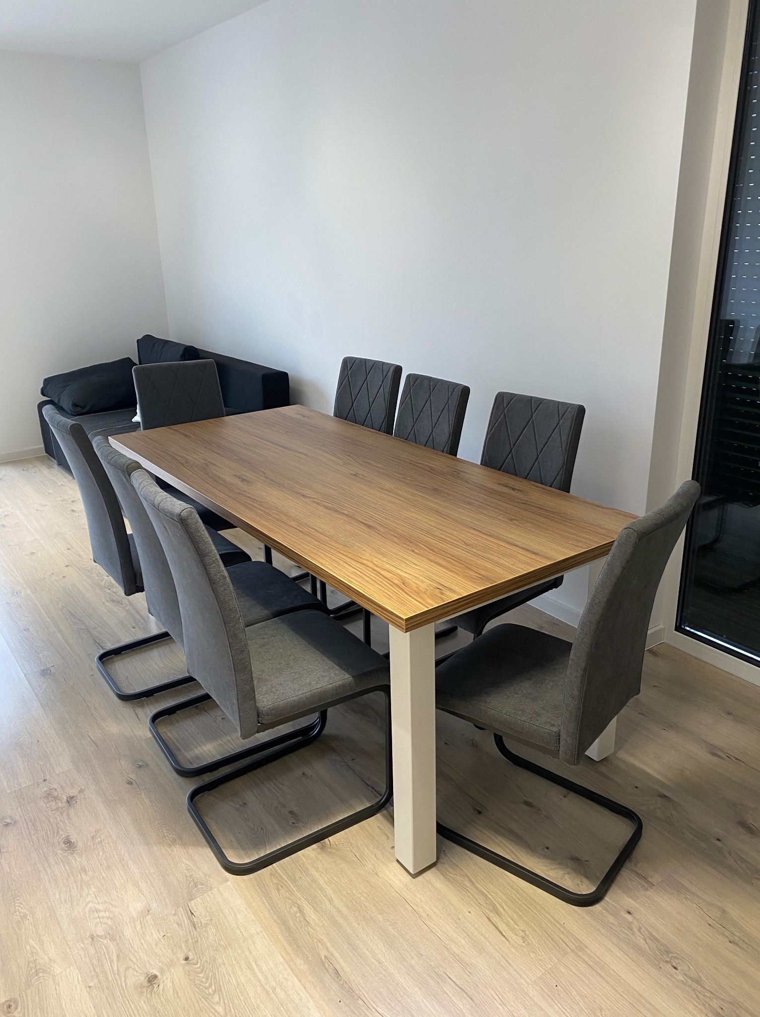 Stół z blatem imitującym drewno i białymi nogami, otoczony sześcioma szarymi krzesłami z czarnymi nogami, ustawiony w minimalistycznym wnętrzu z szarą sofą w tle.