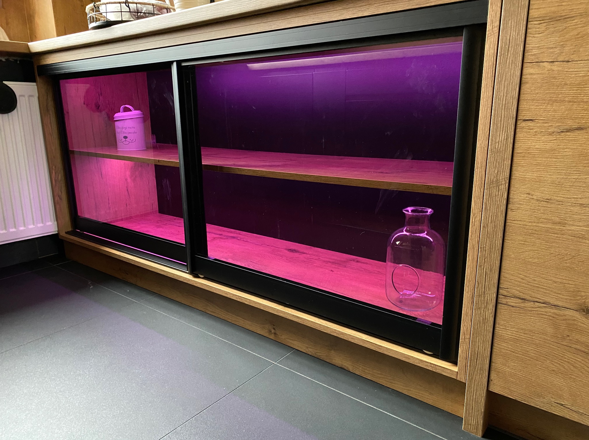 Szafka z przesuwnymi drzwiami, wykonana z drewna w ciepłym odcieniu, oświetlona fioletowym światłem, z dekoracyjnymi przedmiotami na półkach.