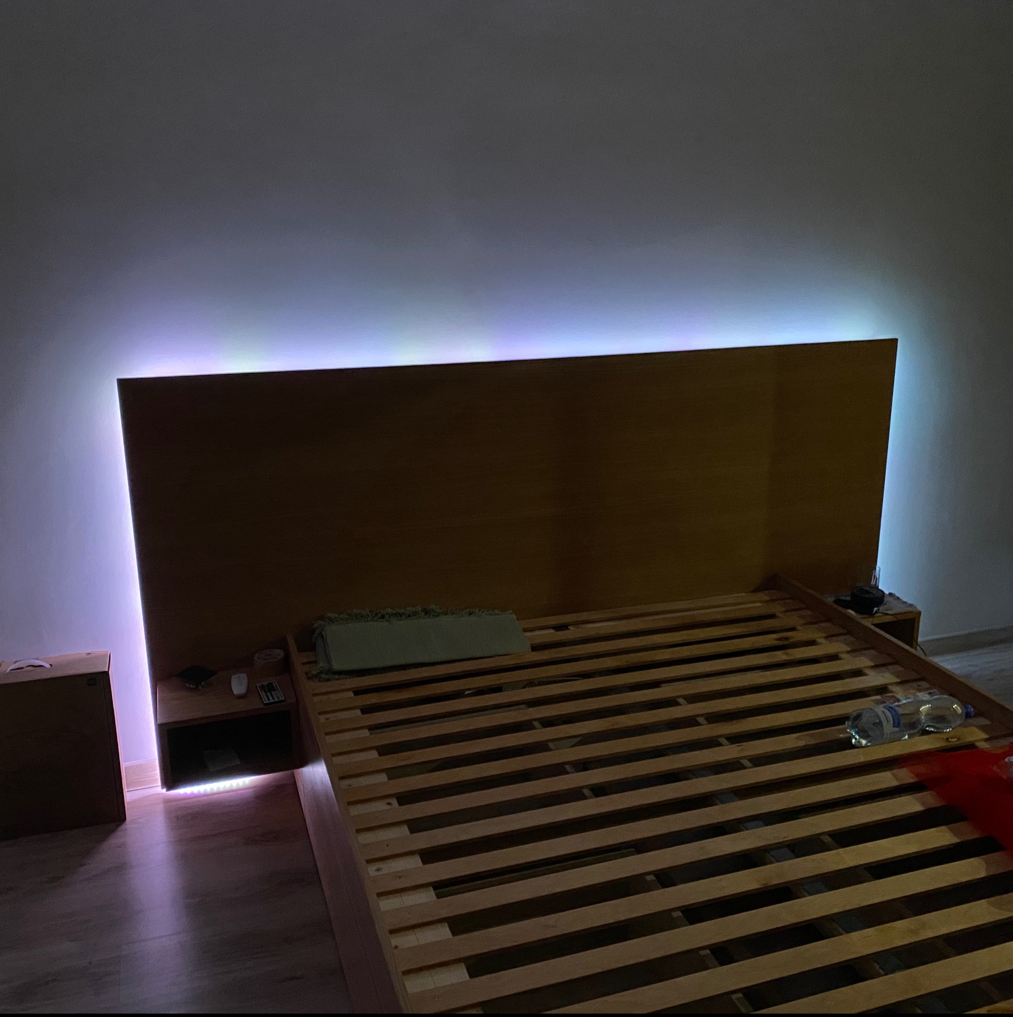 Drewniana rama łóżka z podświetleniem LED, widoczne szafki nocne z akcesoriami i butelka wody na stelażu łóżka.