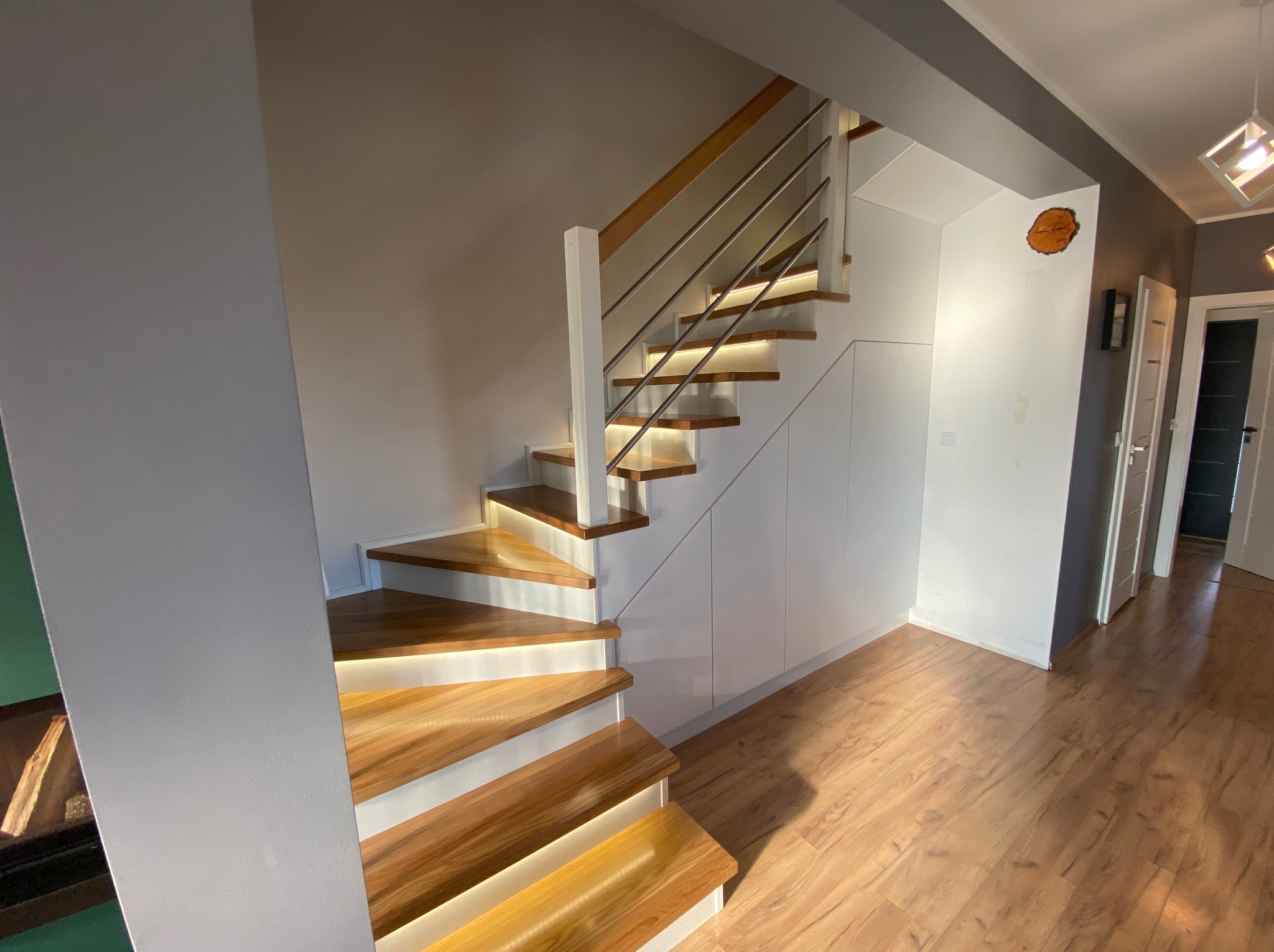 Nowoczesne schody z podświetlanymi stopniami, drewniane stopnie i białe podstopnice, balustrada ze stali nierdzewnej, zabudowa pod schodami z białymi frontami, drewniana podłoga.