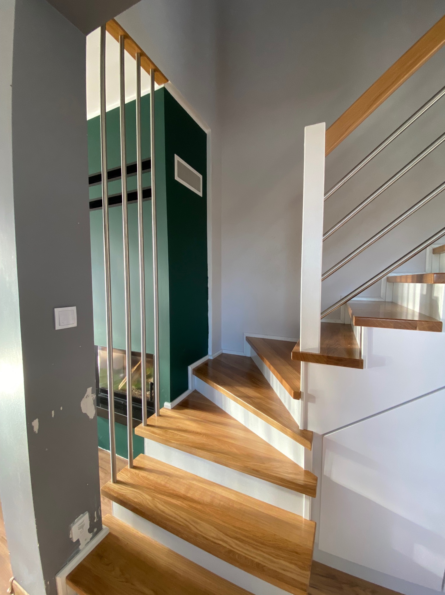 Nowoczesne schody z drewnianymi stopniami, białymi podstopnicami i metalowymi balustradami, widok z dołu, ściana w tle szara z widocznymi śladami po naprawach, zielona ściana w tle.