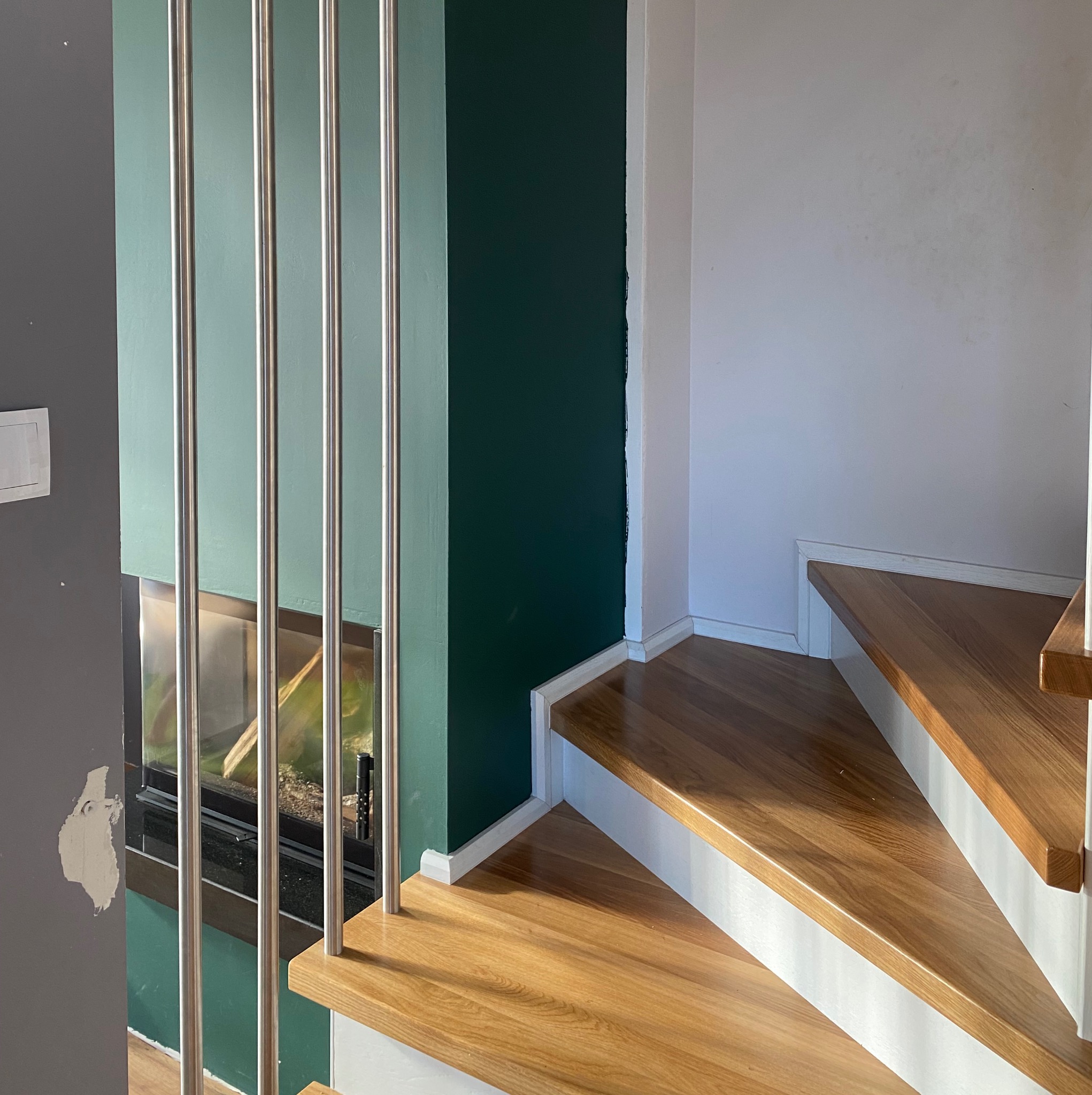 Fragment drewnianych schodów z metalową balustradą na tle ściany w kolorze butelkowej zieleni i akwarium.