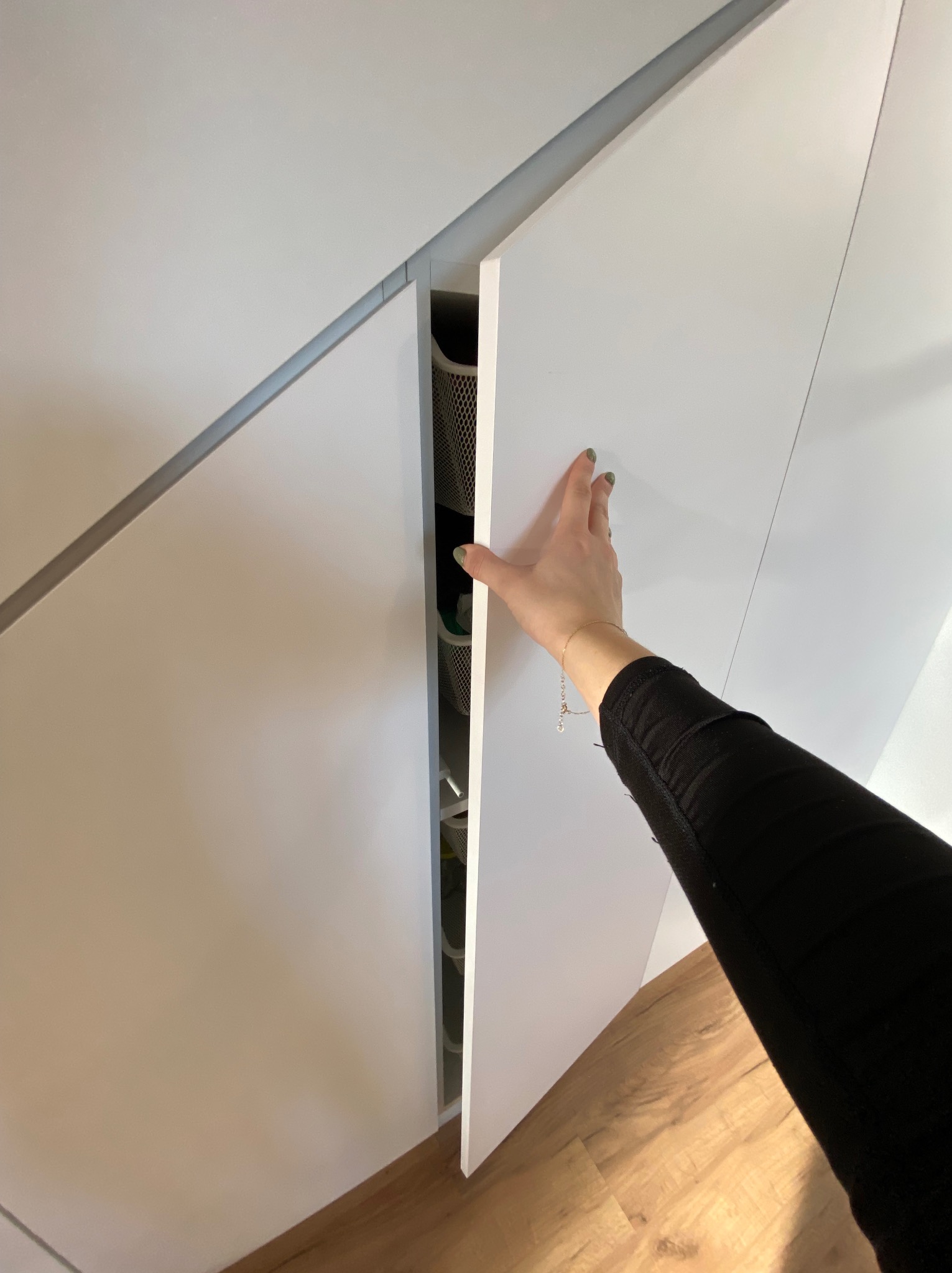 Biała szafa wnękowa z szufladami, częściowo otwarta przez kobiecą dłoń z bransoletką, widoczny parkiet na podłodze.