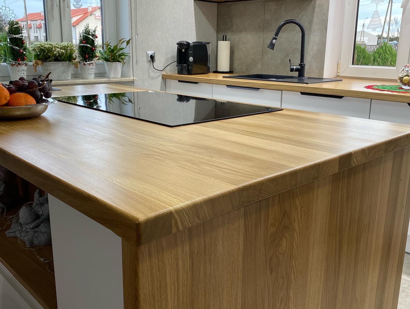 Jasny, drewniany blat kuchenny z wbudowaną płytą indukcyjną, widoczne owoce w misie, okno z roślinami i dekoracjami świątecznymi w tle.