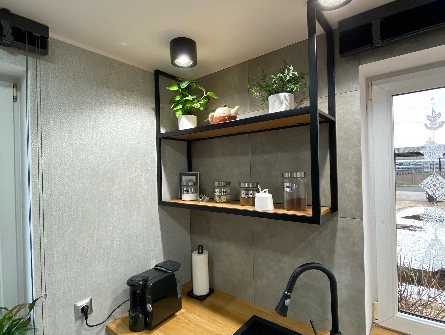 Półka kuchenna z drewnianymi blatami i czarną metalową ramą, na której stoją przyprawy w słoikach, doniczki z roślinami i czajnik, z widocznym ekspresem do kawy na blacie.