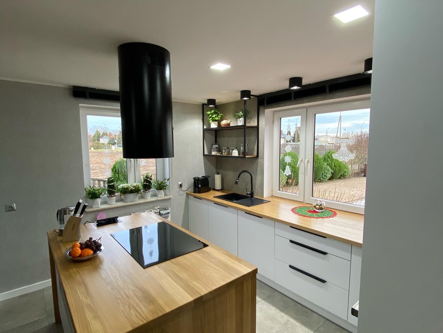 Nowoczesna kuchnia z wyspą: białe szafki z drewnianym blatem, czarna armatura i okap, wyspa z płytą indukcyjną, widok na ogród z dekoracją okienną.