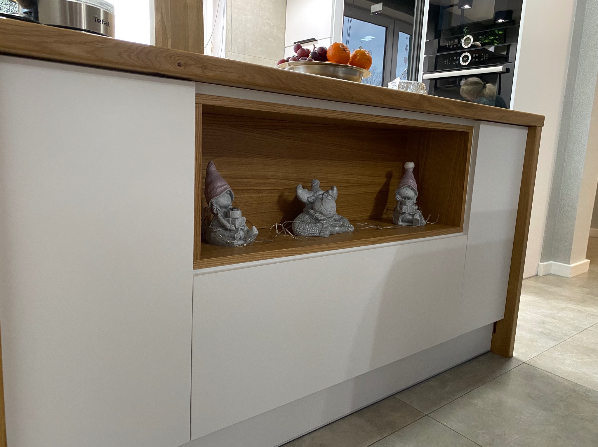 Biała zabudowa kuchenna z drewnianym blatem i otwartą półką, na której stoją trzy szare figurki dekoracyjne w kształcie krasnali i zwierzęcia.