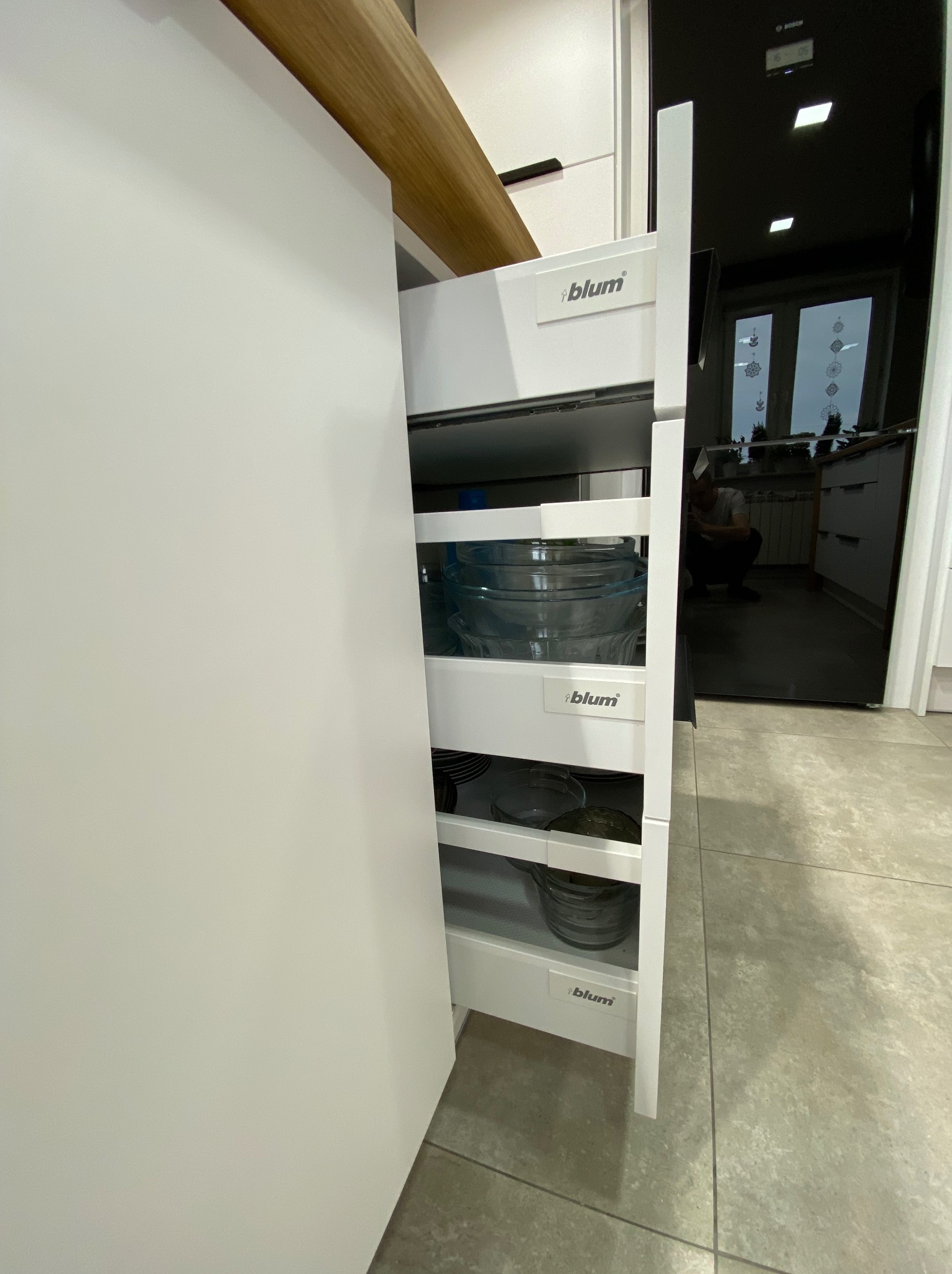 Wysuwana, trzypoziomowa szuflada cargo marki Blum w białej zabudowie kuchennej, wypełniona szklanymi miskami i talerzami.