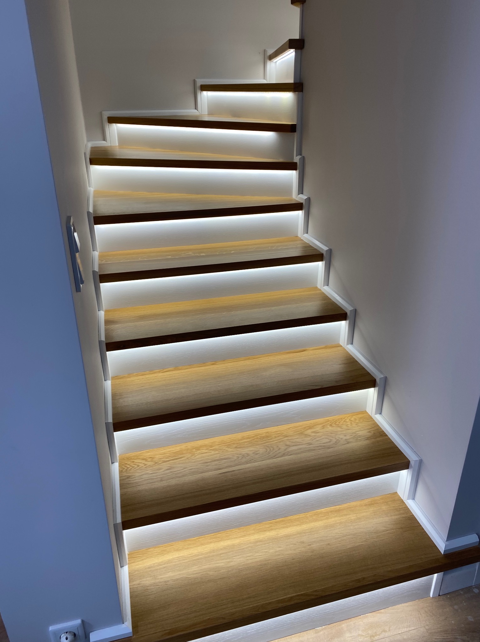 Nowoczesne schody z drewnianymi stopniami i białymi podstopnicami, podświetlone listwami LED, zrealizowane w Śmiglu. Widok z dołu, podkreślający minimalistyczny design i ciepłe światło.
