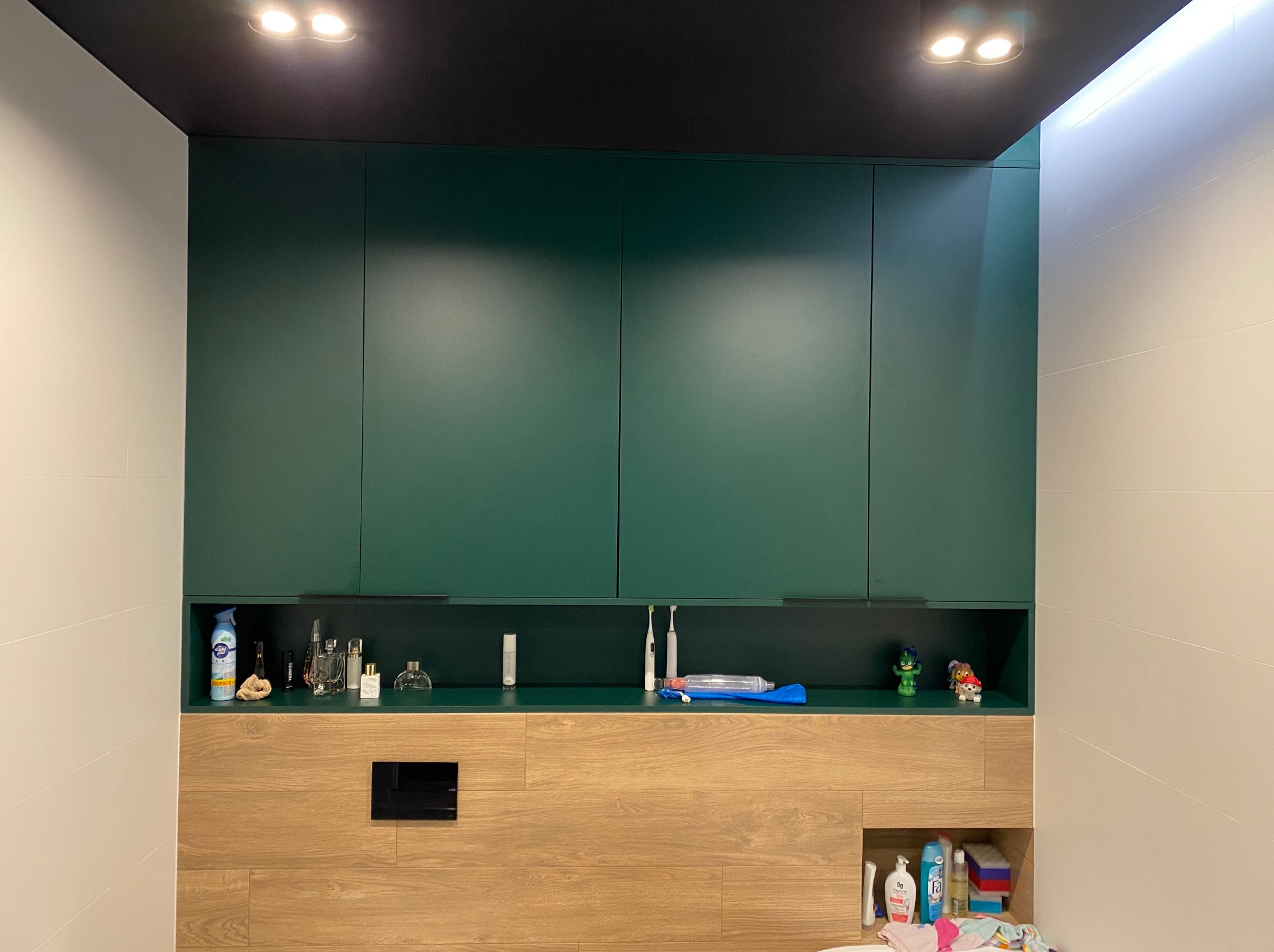 Zielone szafki łazienkowe podwieszane, z otwartą półką na kosmetyki i akcesoria, drewnopodobna zabudowa z ukrytym stelażem WC, białe płytki na ścianach, czarny sufit z oświetleniem punktowym.