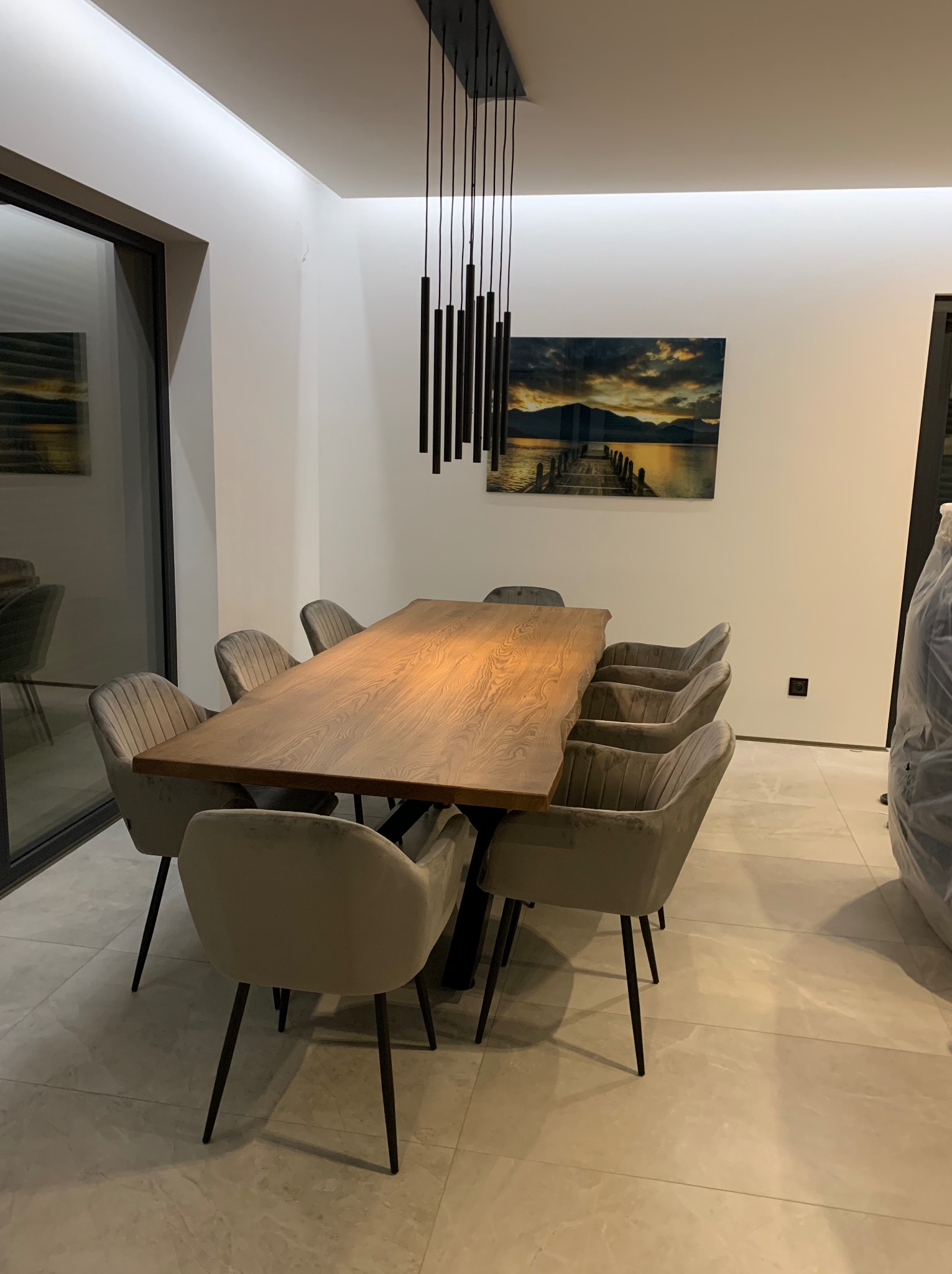 Stół z blatem z litego drewna o nieregularnych krawędziach, otoczony krzesłami tapicerowanymi w odcieniach szarości, oświetlony nowoczesną lampą wiszącą na tle minimalistycznego wnętrza z obrazem.