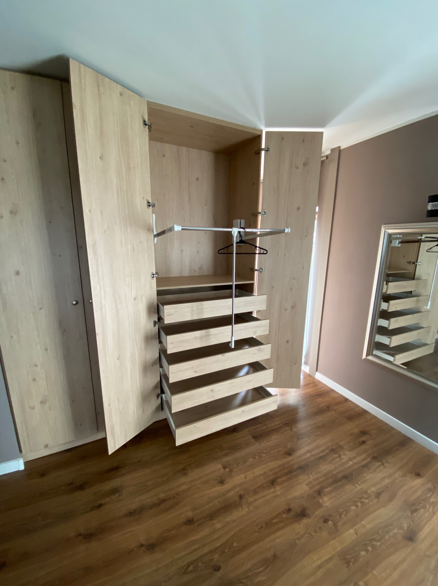 Otwarta szafa wnękowa z jasnego drewna z wysuwanymi szufladami i drążkiem na wieszaki, widoczne zawiasy, brązowa podłoga i ściana w tle.