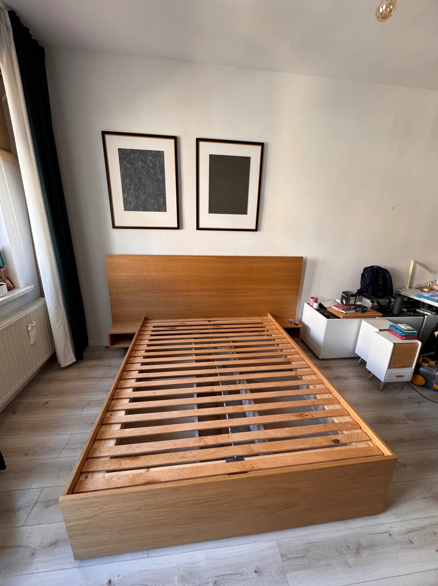 Drewniane łóżko bez materaca z listwowym stelażem, zintegrowane szafki nocne, minimalistyczny design, rama z jasnego drewna, widok z przodu na tle ściany z obrazami.