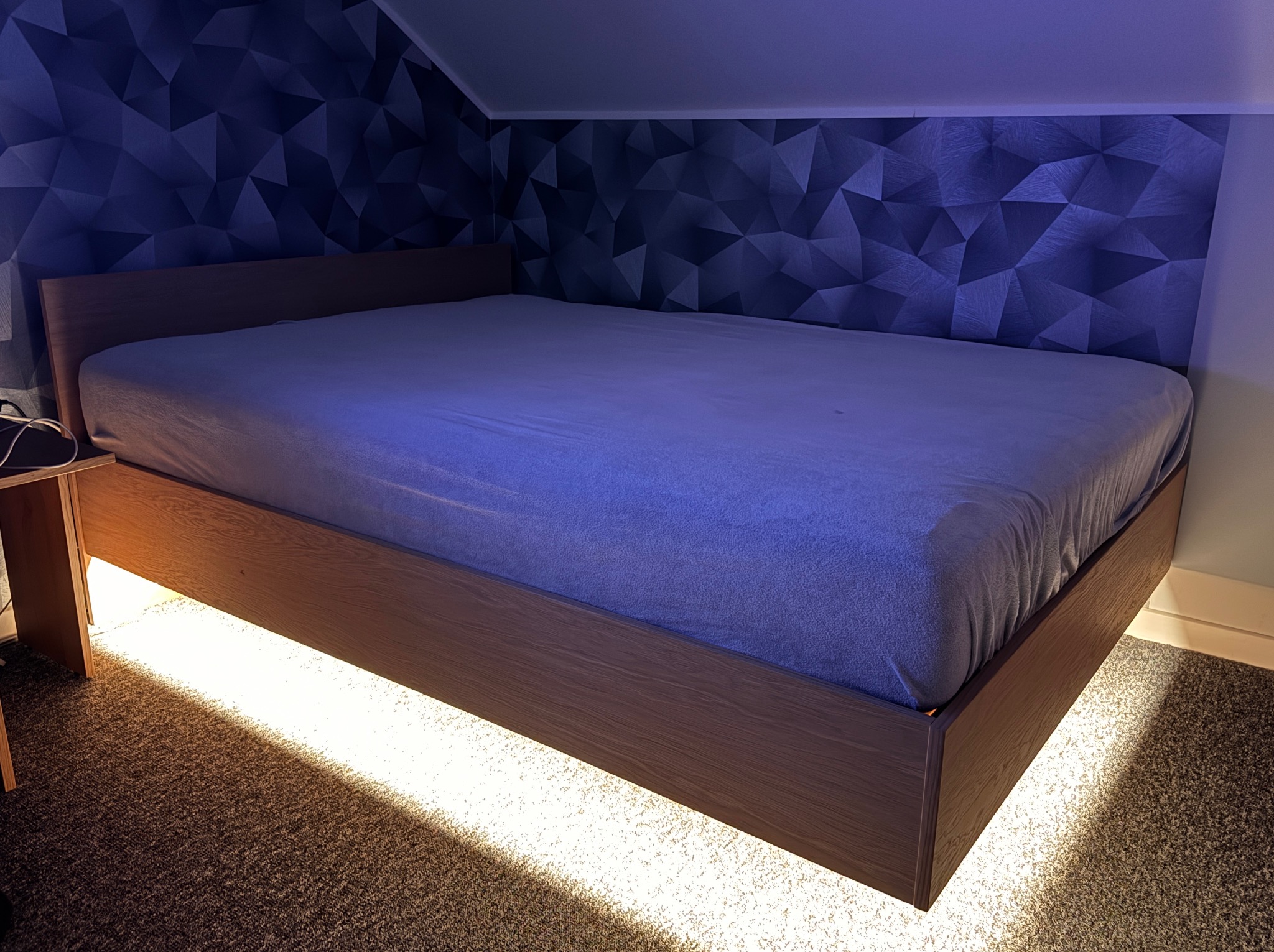 Drewniane łóżko z podświetleniem LED w nowoczesnej sypialni z tapetą w geometryczne wzory. Widoczne oświetlenie pod łóżkiem i stolik nocny po lewej stronie.