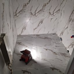 Usługi dekarsko blacharskie Grzegorz Gajewski - Łazienka w trakcie remontu z marmurowymi płytkami na ścianach i podłodze, widoczny grzejnik i narzędzie do docinania płytek.