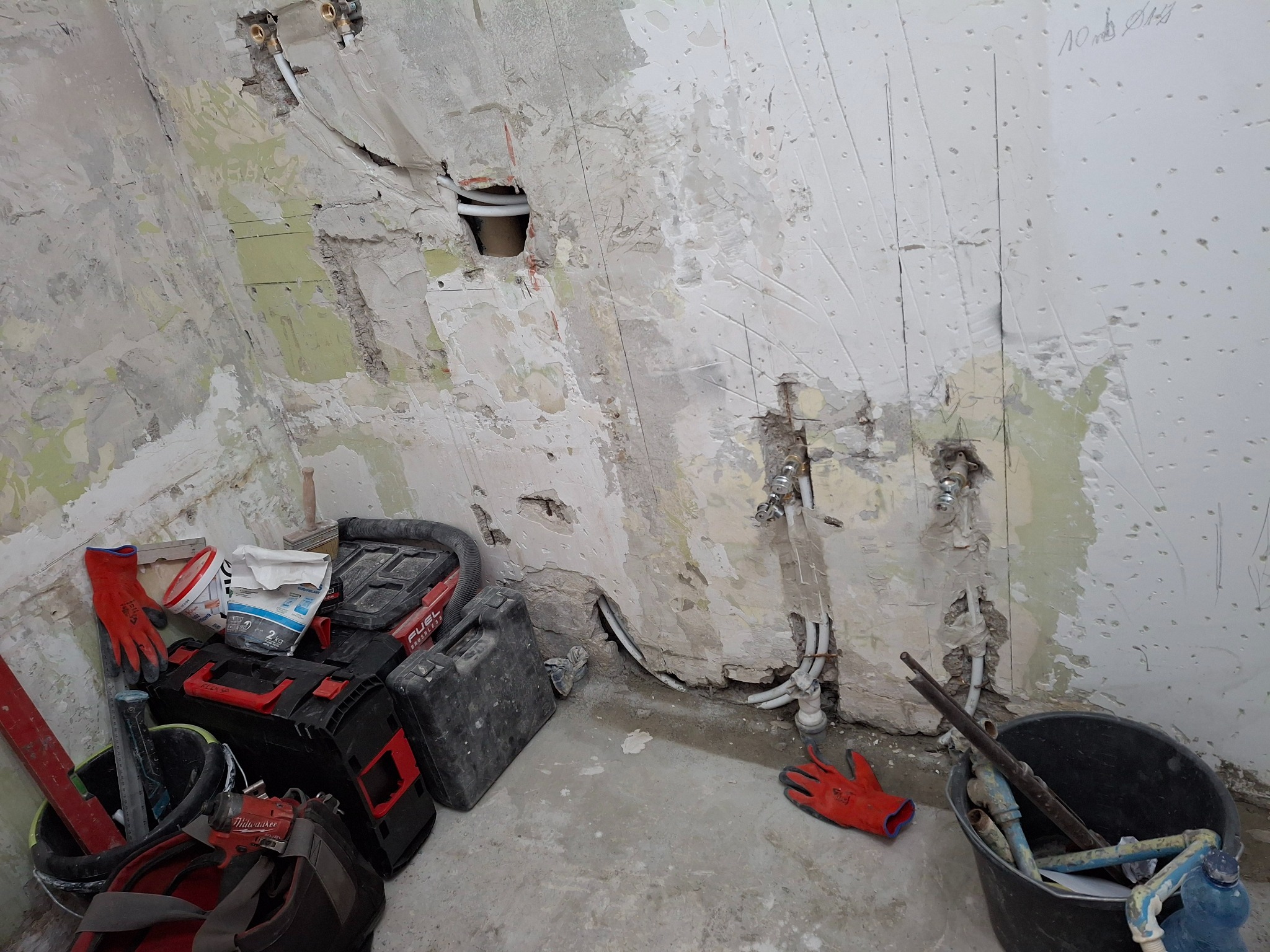 Przygotowanie ściany pod wykończenie w trakcie prac: odsłonięte rury, instalacja wodna, ściana z resztkami starej farby i gipsu, narzędzia w wiadrach i skrzynkach, czerwone rękawice robocze.
