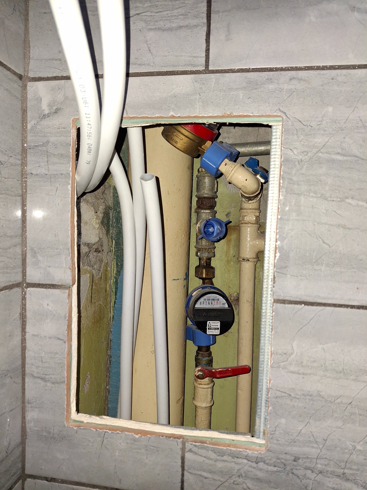 Wnęka w łazience z widocznymi rurami, zaworami i licznikiem wody, otoczona szarą płytką imitującą marmur.