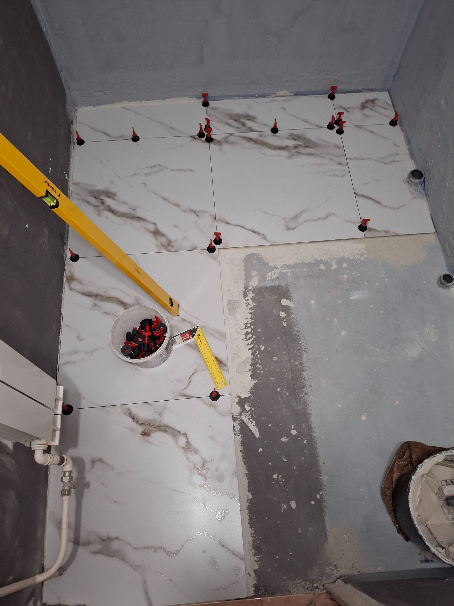 Układanie płytek imitujących marmur na podłodze w łazience z użyciem poziomicy, systemu poziomowania płytek i pojemnika z klinami.