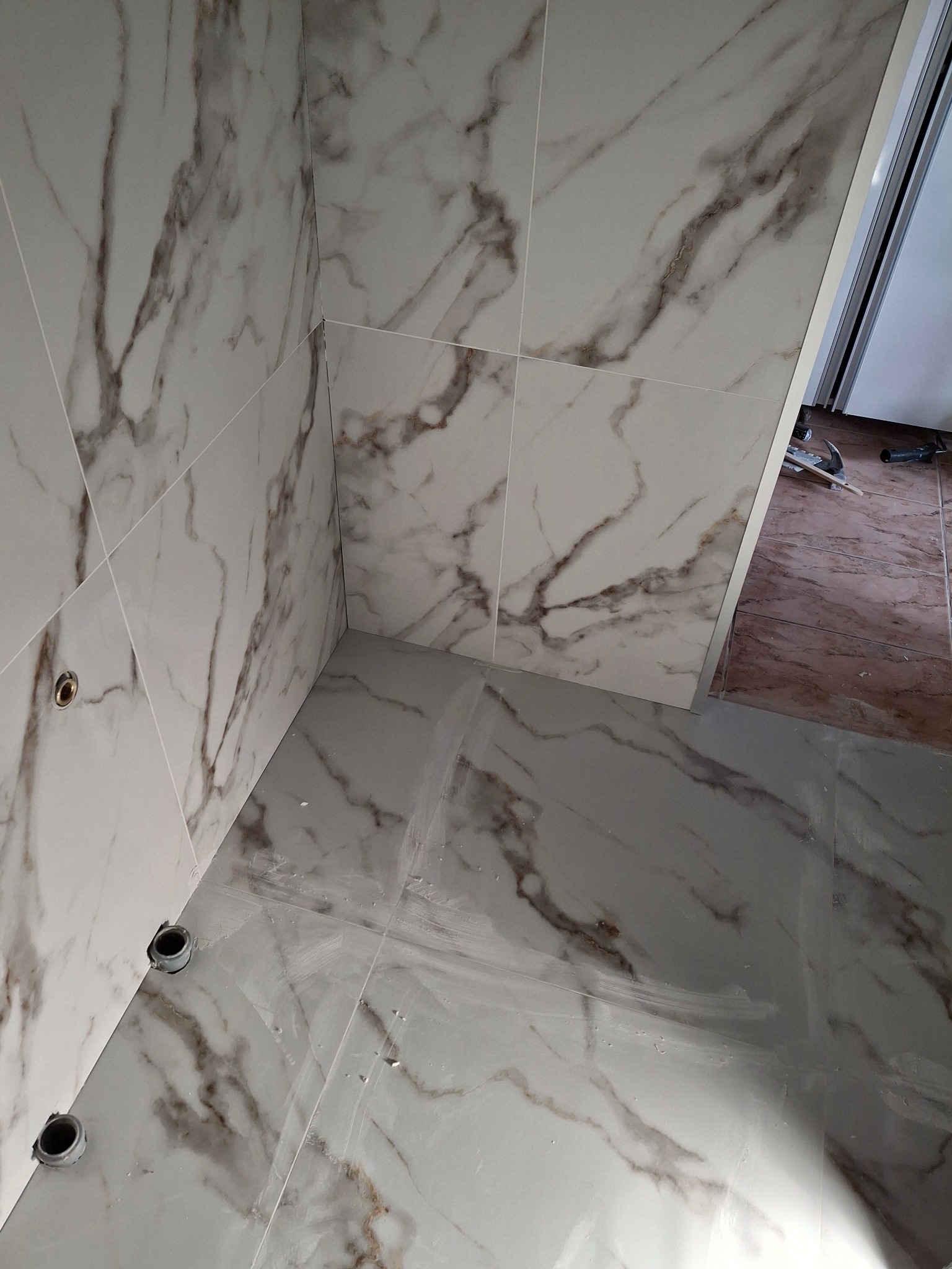Fragment narożnika łazienki z białymi płytkami imitującymi marmur na ścianach i podłodze, widoczne otwory na instalacje, częściowo widoczne narzędzia na podłodze w tle.