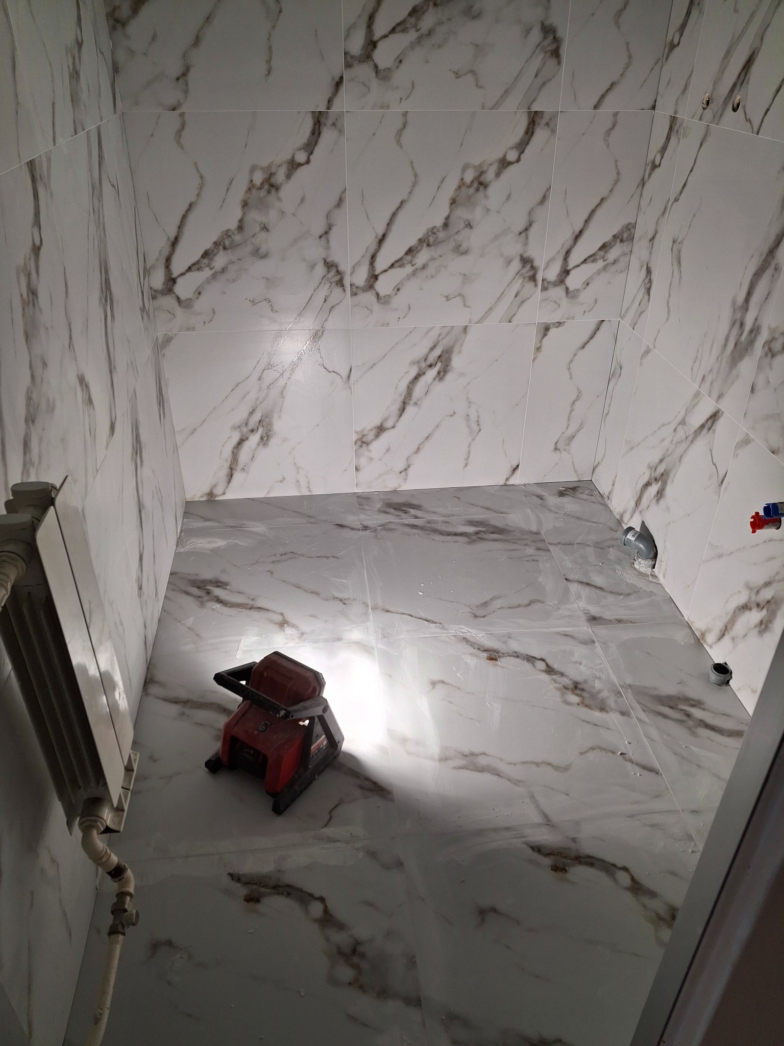 Łazienka w trakcie remontu z marmurowymi płytkami na ścianach i podłodze, widoczny grzejnik i narzędzie do docinania płytek.
