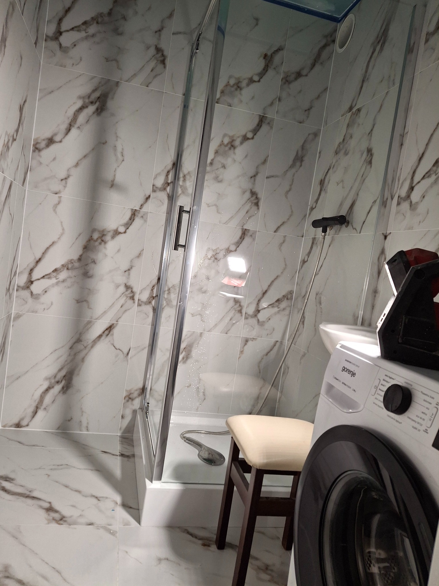 Nowoczesna kabina prysznicowa z białymi płytkami imitującymi marmur, widoczna pralka Gorenje i stołek w tle, elementy instalacji natryskowej.