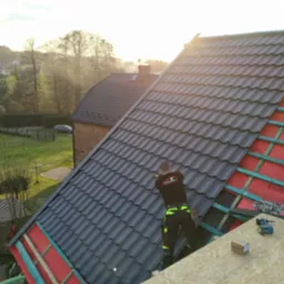 Dekarz montuje grafitową dach&oacute;wkę na nowym dachu, widoczne narzędzia i elementy konstrukcji dachu na tle zabudowań i zieleni.