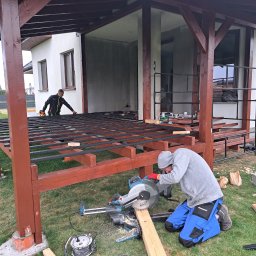 Wojciech Bejcar Dachcover - Budowa drewnianej konstrukcji z metalowymi elementami. Pracownik tnie deskę piłą elektryczną, drugi montuje elementy na górze konstrukcji. Widoczne narzędzia i trawa.