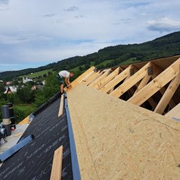 Wojciech Bejcar Dachcover - Pracownik montuje płyty OSB na drewnianej konstrukcji dachu, widok na zielone wzgórza w tle. Czarna membrana dachowa z widocznym napisem TOPDEK.