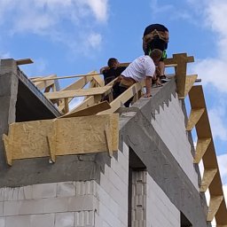 Wojciech Bejcar Dachcover - Ekipa monterów na szczycie budynku w trakcie budowy drewnianej konstrukcji dachu na tle błękitnego nieba z chmurami. Widoczne narzędzia i elementy konstrukcyjne.