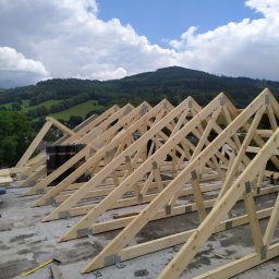 Wojciech Bejcar Dachcover - Wznoszenie drewnianej więźby dachowej na betonowej płycie, widok na zalesione wzgórze pod błękitnym niebem z chmurami. Elementy złączone metalowymi płytkami.