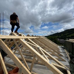 Wojciech Bejcar Dachcover - Monter z narzędziami na drewnianej konstrukcji dachu na tle pochmurnego nieba i zielonych wzgórz. Widoczne elementy mocujące i cegły.