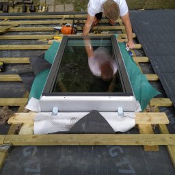 Wojciech Bejcar Dachcover - Montaż okna dachowego na drewnianej konstrukcji dachu, widok z góry. Pracownik w koszulce z logo firmy dekarskiej. Widoczne narzędzia i materiały izolacyjne.