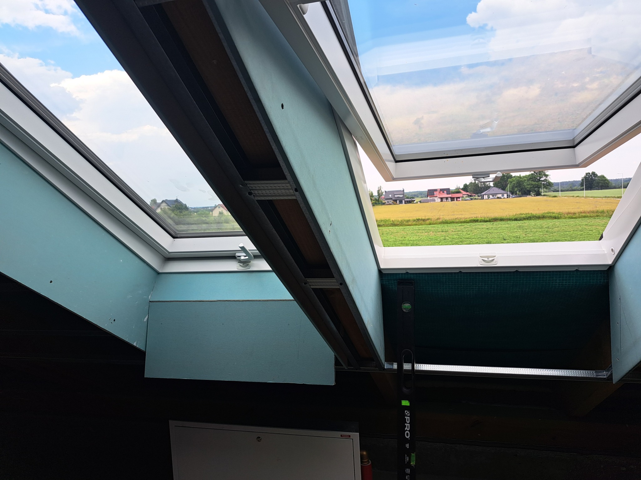 Montaż okien dachowych w poddaszu z widokiem na wiejski krajobraz, widoczne elementy konstrukcyjne i poziomica.