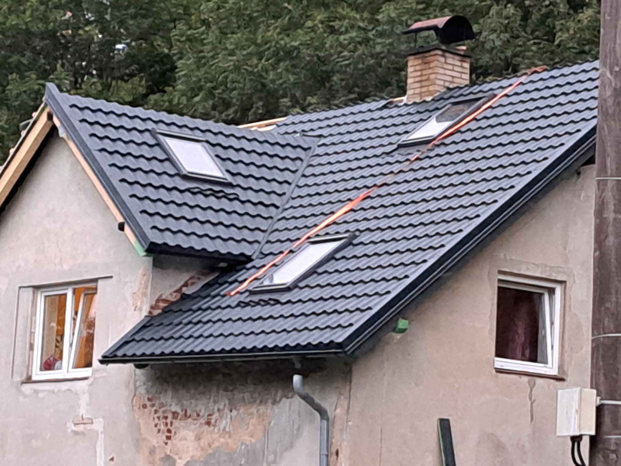 Dom z nowym, ciemnoszarym dachem z blachy trapezowej z dwoma oknami dachowymi, widoczne fragmenty starej elewacji i nowej więźby dachowej.