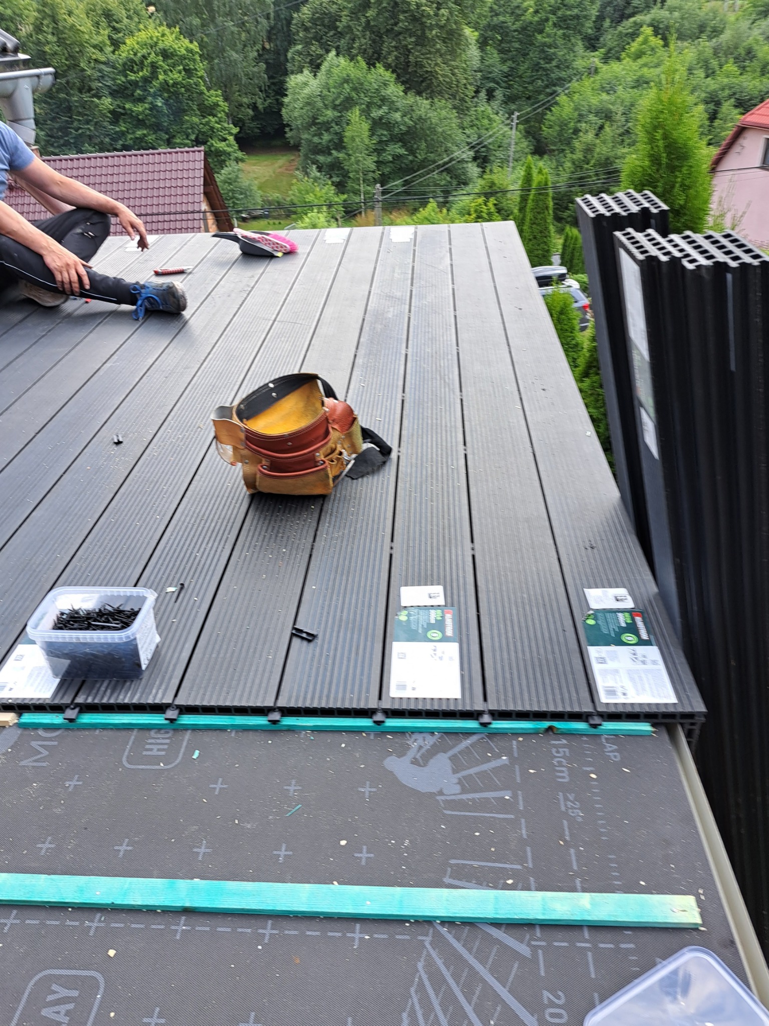 Montaż kompozytowej deski tarasowej na dachu, widoczny pas narzędzi, pojemnik z wkrętami, membrana dachowa z nadrukiem, oraz fragment sylwetki dekarza w tle.