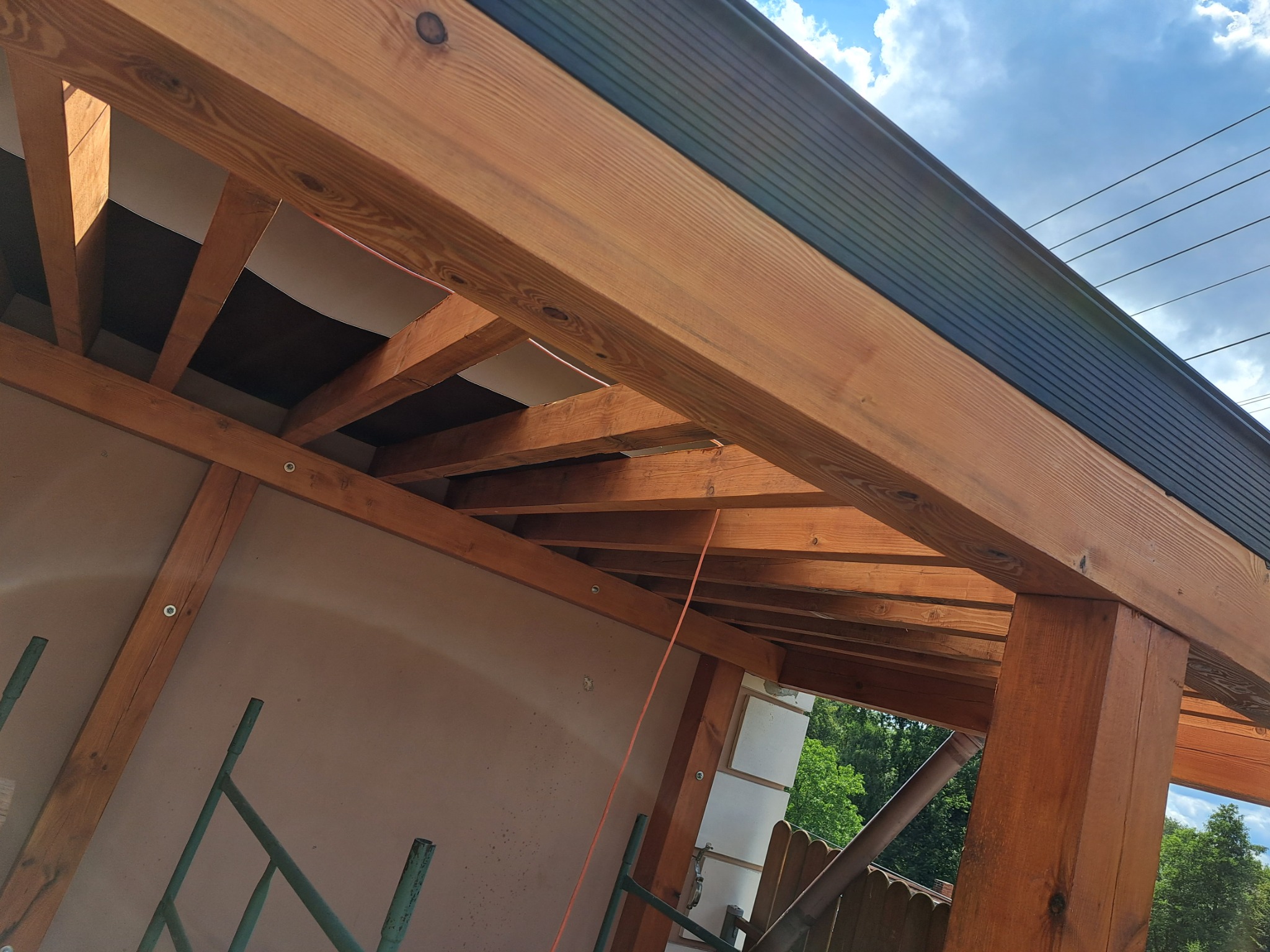 Wiązar dachowy z belek z widoczną izolacją i fragmentem poszycia dachu od dołu, z elementami rusztowania i niebem w tle.