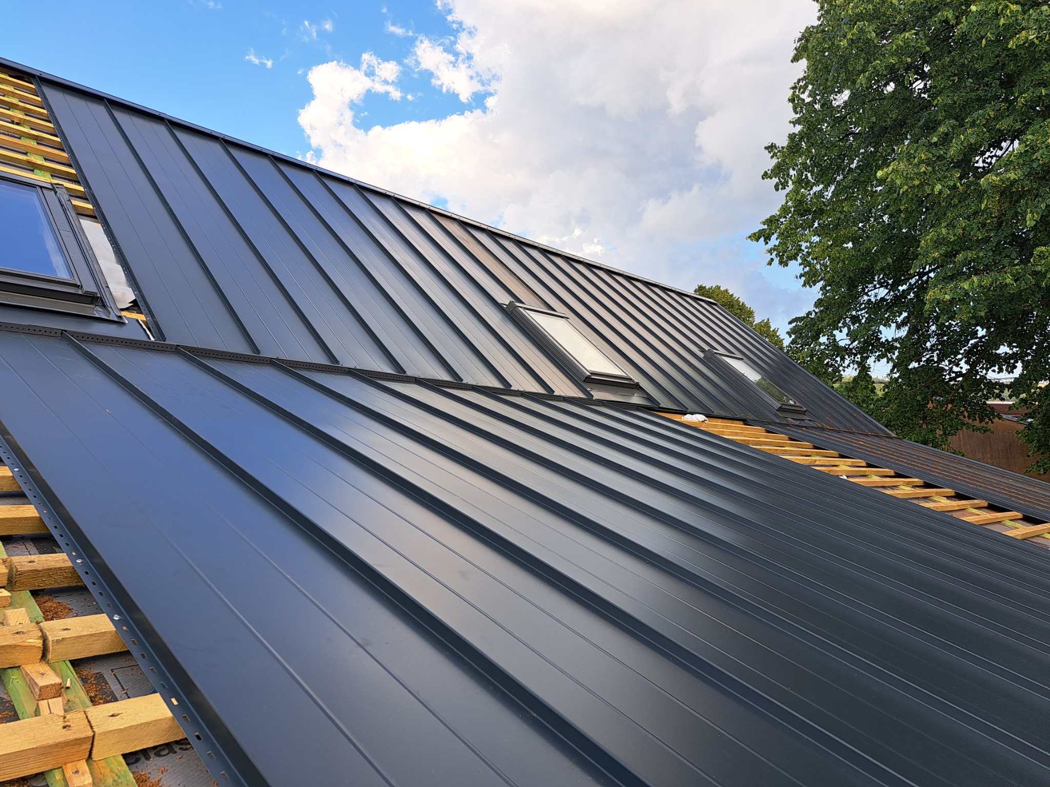 Nowe, grafitowe pokrycie dachu z blachy trapezowej z oknami dachowymi, widoczne drewniane łaty i fragment starego pokrycia, błękitne niebo z chmurami w tle.